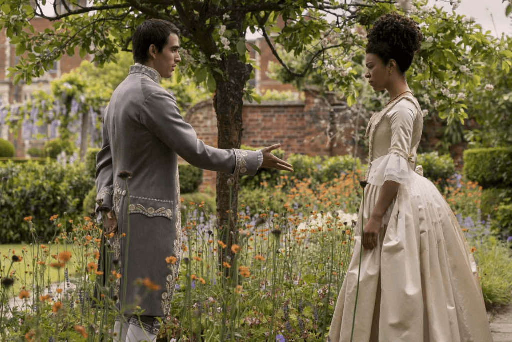 Cena da série Rainha Charlotte, mostrando George e Charlotte em um jardim repleto de flores coloridas. George, vestido com um traje cinza bordado, estende a mão em um gesto convidativo. Charlotte, usando um vestido bege elegante com detalhes clássicos, observa a mão dele com uma expressão séria. O cenário inclui uma árvore sombreando os personagens e um fundo com vegetação vibrante e muros de tijolos.